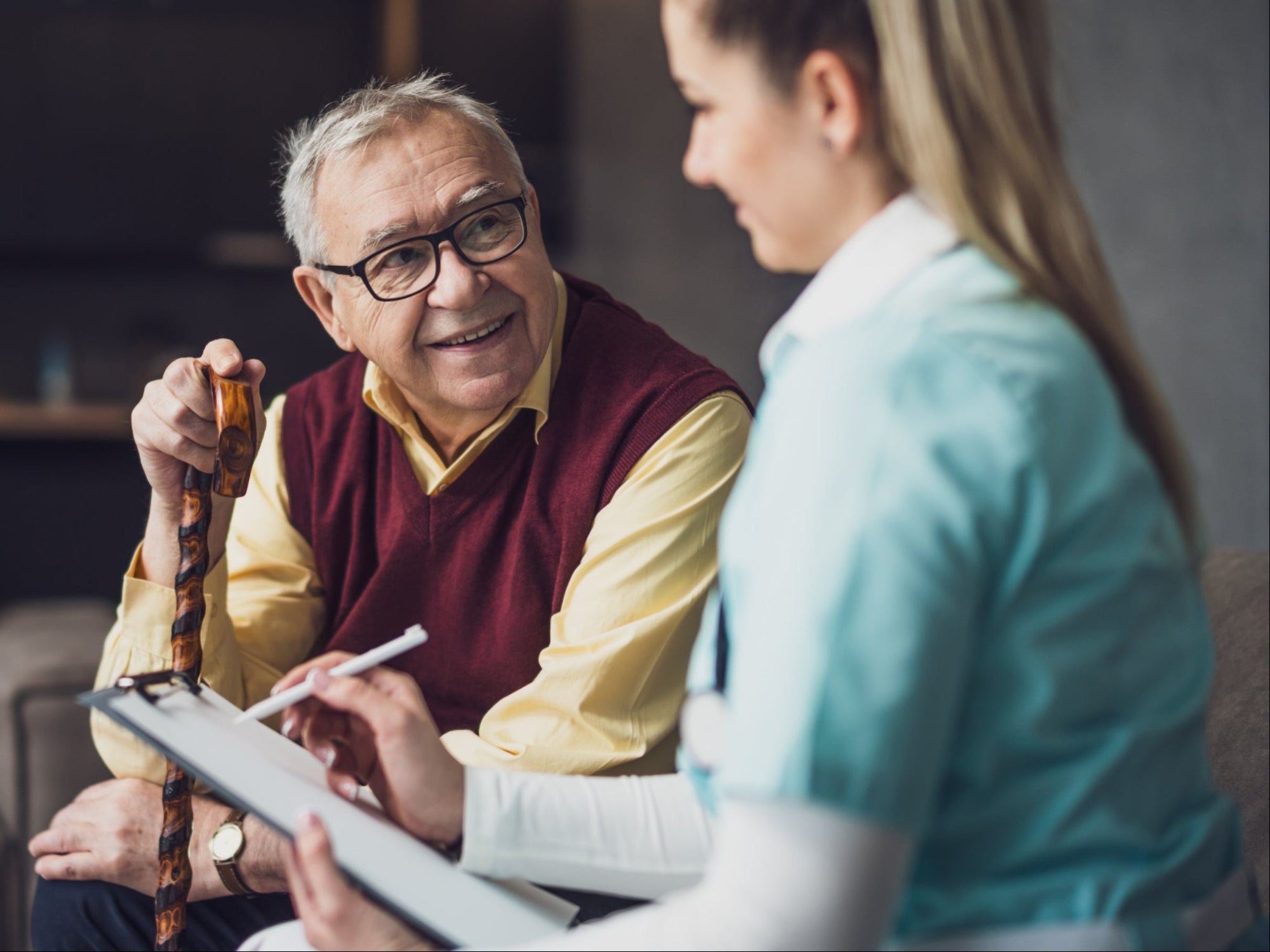 An elderly man wearing glasses and a maroon sweater vest over a yellow shirt is sitting and talking with a female healthcare provider in a teal uniform, holding a clipboard and cane.